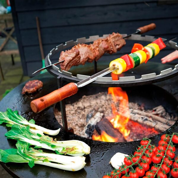 Brochetas de carne y verduras cocinándose sobre el fuego en un set OFYR, con vegetales frescos a un lado.