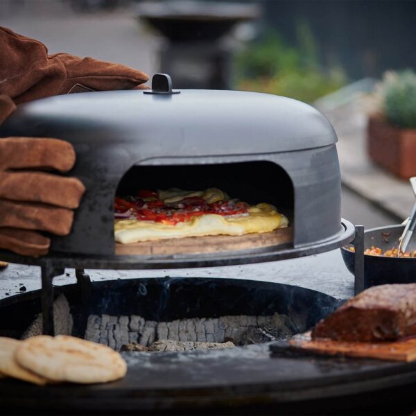 Horno de pizza OFYR con una pizza cocinándose bajo el cloche, mientras se usan guantes resistentes al calor para ajustar el horno y cocinar a leña al aire libre.