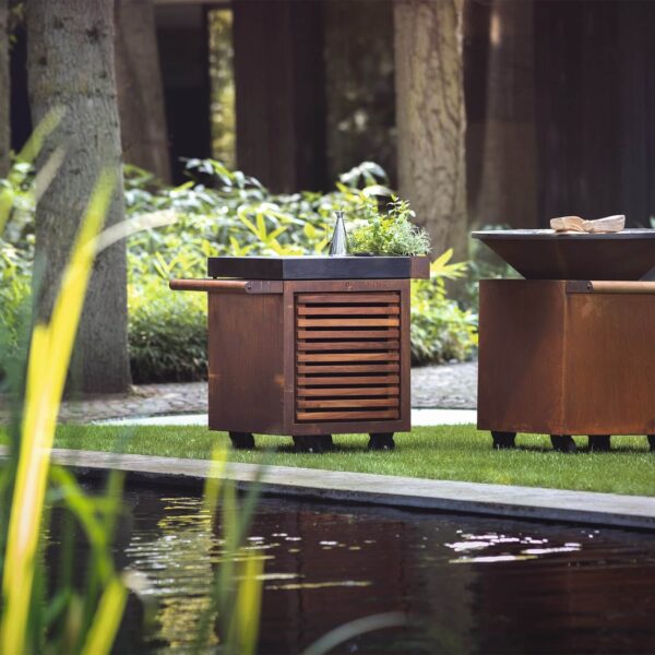 Mesa OFYR Kamado de madera oscura en un jardín junto a un estanque, utilizada como estación de cocina al aire libre.