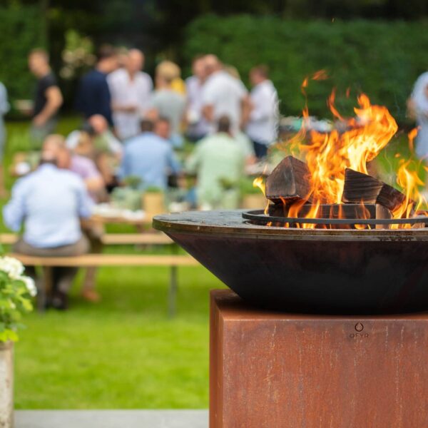 Barbacoa de leña OFYR encendida en un jardín con personas disfrutando de una reunión al fondo.