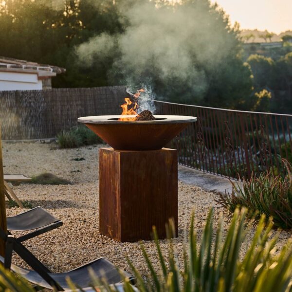 Barbacoa de leña OFYR en acero Corten encendida en un jardín al atardecer.