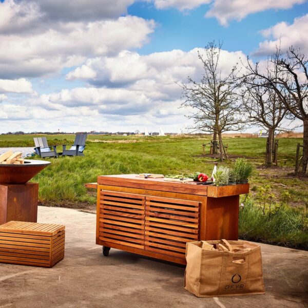 Mesa OFYR Kamado en un entorno campestre, equipada con una superficie de trabajo de madera y accesorios de cocina al aire libre.