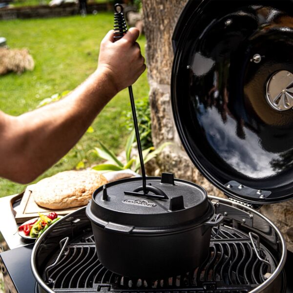 Persona levantando la tapa de una olla holandesa de hierro fundido con un gancho especial, sobre una parrilla al aire libre.