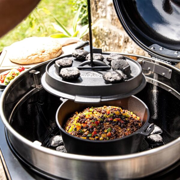 Guiso cocinándose en olla holandesa de hierro fundido bajo brasas calientes en un entorno de barbacoa al aire libre.