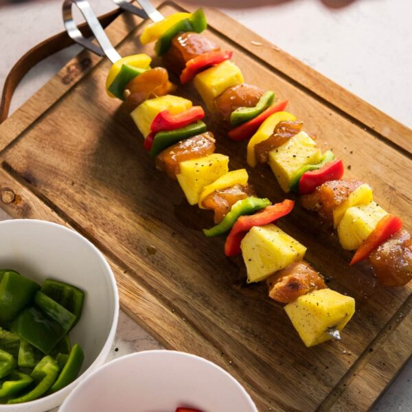 Brochetas de acero inoxidable con trozos de pollo, piña y pimientos de colores colocadas sobre una tabla de madera.