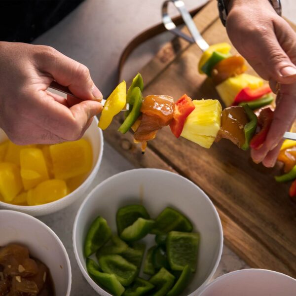 Persona preparando brochetas con trozos de pollo, piña y pimientos de colores, utilizando brochetas de acero inoxidable sobre una tabla de madera.