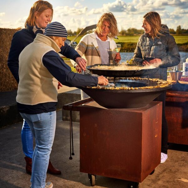 Grupo de personas cocinando juntas en una Barbacoa de Leña Classic Pro en acabado corten durante una reunión al aire libre.