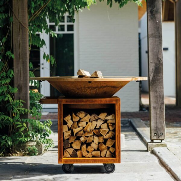 Barbacoa de Leña Classic Pro en acabado corten con almacenamiento de leña, ubicada en un jardín exterior.