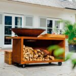 Barbacoa OFYR Island 100 PRO de acero corten en un patio, con almacenamiento de leña y encimera de teca, lista para cocinar al aire libre.