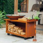 Barbacoa OFYR Island 100 PRO de acero corten en un jardín, con almacenamiento de leña, encimera de teca y herramientas de parrilla al lado