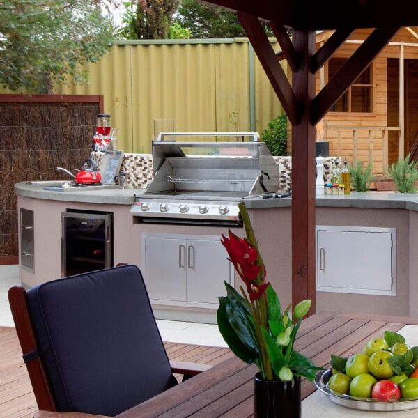 Cocina al aire libre con barbacoa de acero inoxidable, horno de leña y área de comedor bajo un gazebo de madera.