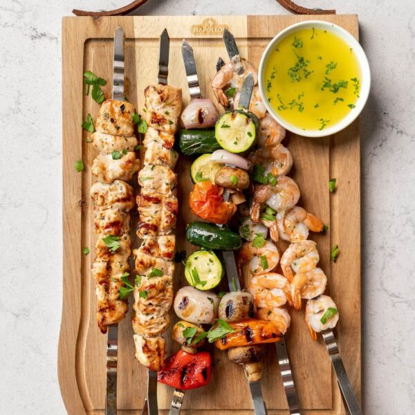 Brochetas de acero inoxidable con pollo, camarones y vegetales variados, servidas en una tabla de madera junto a un recipiente de salsa de hierbas.