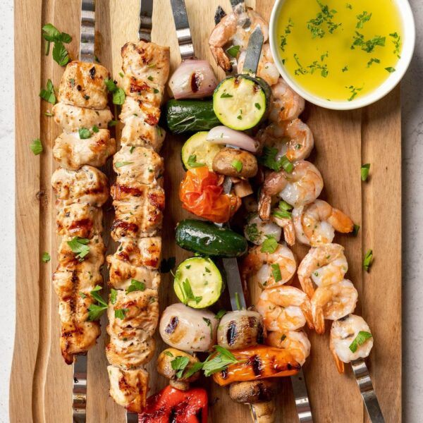 Brochetas de acero inoxidable con pollo, camarones, vegetales y champiñones, servidas en una tabla de madera con un recipiente de salsa de hierbas.