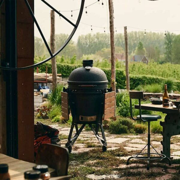 Kamado Urban Large 49 cm en un jardín con mesas auxiliares plegadas y carrito de acero, rodeado de vegetación y luces colgantes.