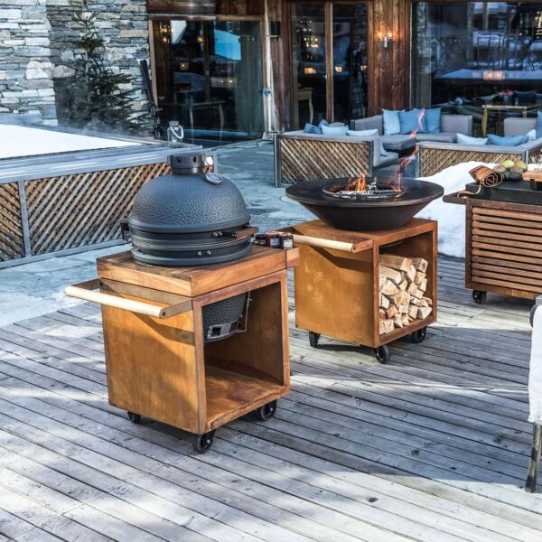 Kamado Urban Large 49 cm sobre una mesa de madera con ruedas, en un entorno exterior con chimenea y decoración de madera en una terraza de montaña.