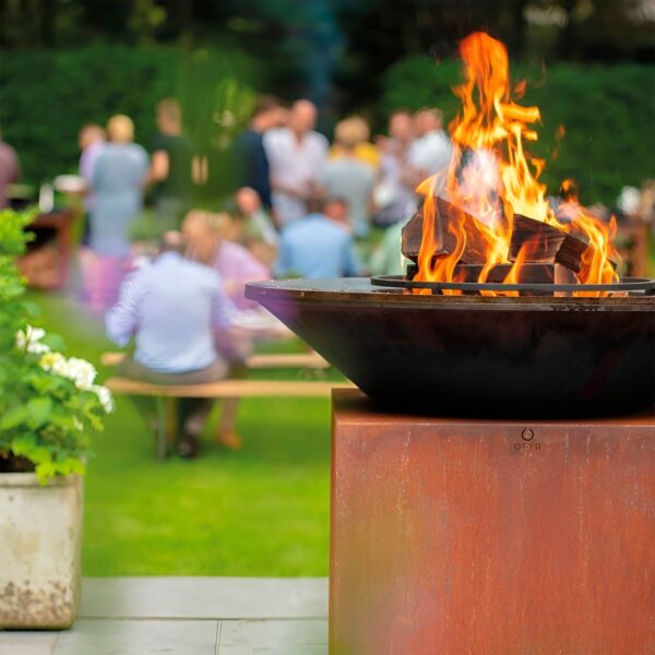 Barbacoa OFYR encendida en un jardín con personas en el fondo disfrutando de una reunión al aire libre.