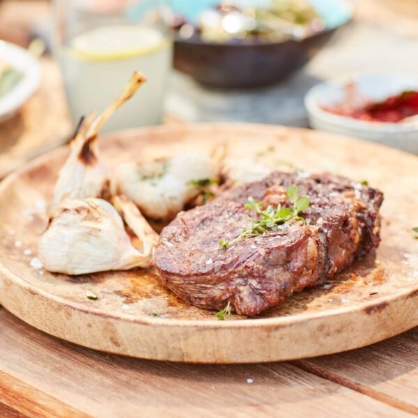 Bistec asado servido en un plato de madera con ajos asados y hierbas frescas.