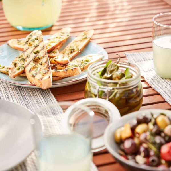 Panes tostados con hierbas servidos en un plato junto a un frasco de pimientos y un tazón de aceitunas.