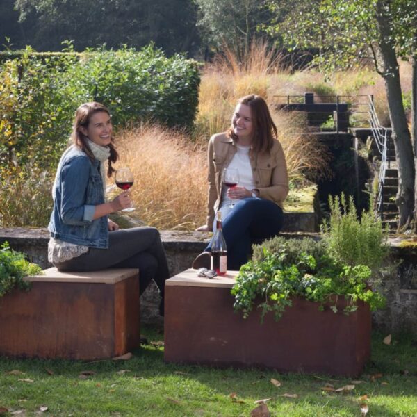 Dos mujeres sentadas en bancos de jardín de acero corten con jardineras integradas, disfrutando de una copa de vino en un espacio exterior.