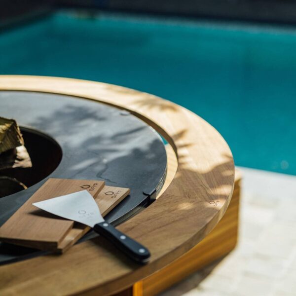 Detalle del Anillo de Protección Contra Quemaduras de madera de teca en barbacoa OFYR con utensilios junto a la piscina.