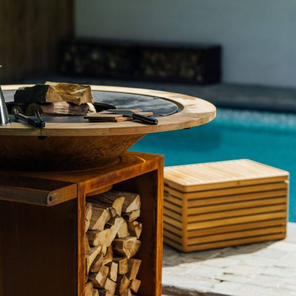 Barbacoa OFYR con Anillo de Protección Contra Quemaduras de madera de teca y almacenamiento de leña junto a una piscina.