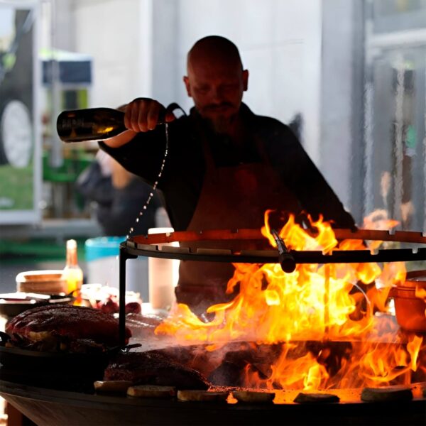 Cocinero añadiendo líquido sobre un set OFYR con grandes llamas mientras cocina carne al fuego.