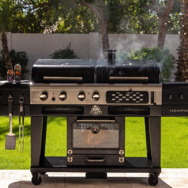 Barbacoa Pit Boss Memphis V2 en uso, emitiendo humo desde su parrilla de carbón y ahumador eléctrico, con utensilios y salsas en un jardín al aire libre.