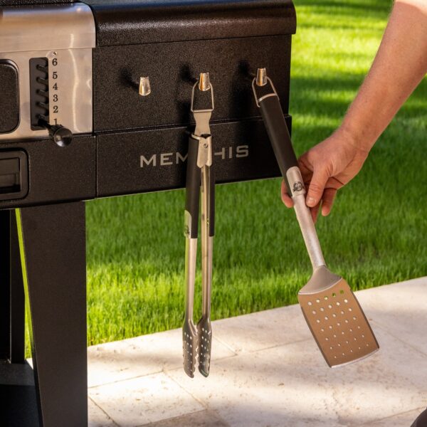 Persona colgando una espátula en los ganchos laterales de la barbacoa Pit Boss Memphis V2, junto a unas pinzas de acero inoxidable.