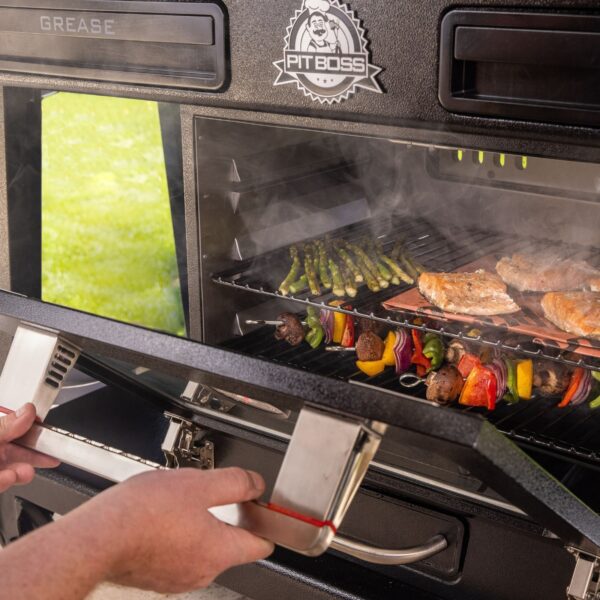 Persona abriendo la puerta del ahumador eléctrico de la Pit Boss Memphis V2, con humo saliendo y alimentos como espárragos, brochetas y filetes de salmón en cocción.