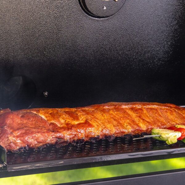 Costillar dorado y jugoso cocinándose en la rejilla superior de la barbacoa Pit Boss Memphis V2, con un ligero ahumado y vegetales a un lado.