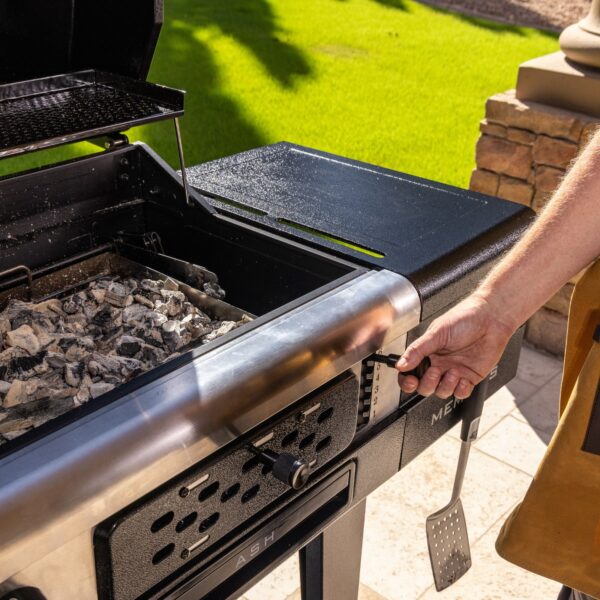 Persona ajustando la altura de la bandeja de carbón en la barbacoa Pit Boss Memphis V2, con la parrilla abierta y brasas encendidas.