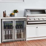 Nevera Beefeater de dos puertas empotrada en una cocina exterior con botellas de vino y cerveza en su interior, junto a una parrilla de acero inoxidable.