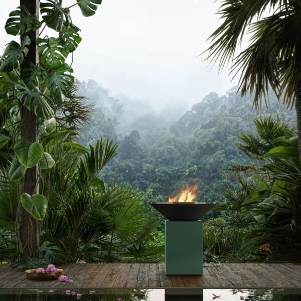 Barbacoa OFYR Chrome Green con fuego encendido en una terraza de madera rodeada de selva tropical y vegetación exuberante.