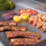 Primer plano de alimentos cocinándose sobre plancha encastrable: costillas, camarones, cebolla, limón y tomate sobre superficie caliente.
