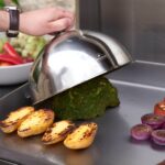 Campana metálica sobre brócoli en plancha encastrable con papas doradas y cebolla cocinándose.