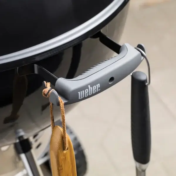 Detalle del asa de la Weber Original Kettle Premium con logotipo Weber y un guante colgado del gancho portautensilios.