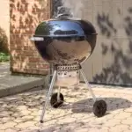 Barbacoa Weber Original Kettle Premium de 57 cm en un patio exterior junto a un muro de ladrillo.