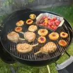 Chuletas, mitades de melocotón y tomates cocinándose en una Weber Kettle abierta al aire libre.