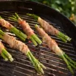 Espárragos envueltos en bacon cocinándose sobre la parrilla de carbón de una Weber Kettle.