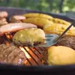 Hamburguesas con queso, salchichas y mazorcas cocinándose en una Weber Kettle mientras una espátula levanta una hamburguesa.