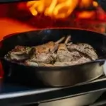 Chuletas de cordero dorándose en una sartén de hierro dentro de un horno con la llama al fondo.