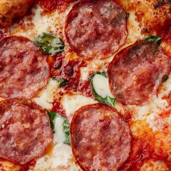 Primer plano de una pizza con pepperoni, albahaca y abundante queso fundido.
