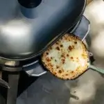 Pizza blanca dorada sobre una pala metálica bajo una tapa de cocción, vista desde arriba.