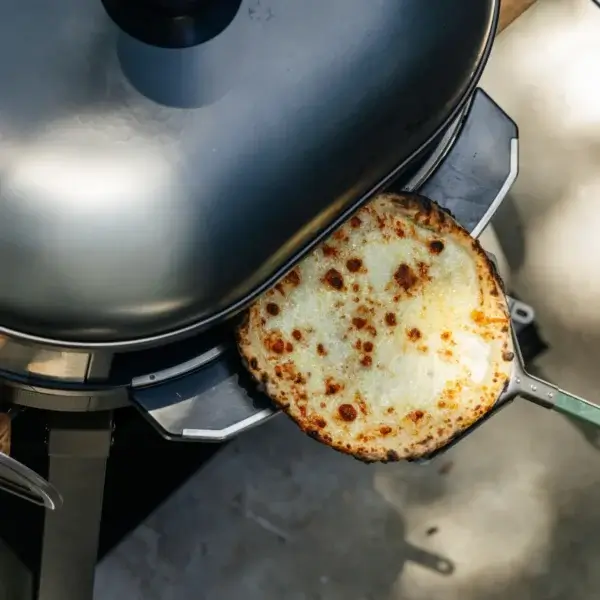 Pizza blanca dorada sobre una pala metálica bajo una tapa de cocción, vista desde arriba.