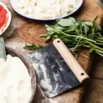 Gozney Dough Cutter con harina sobre la hoja, junto a albahaca, mozzarella y un bol de harina sobre mesa de madera.