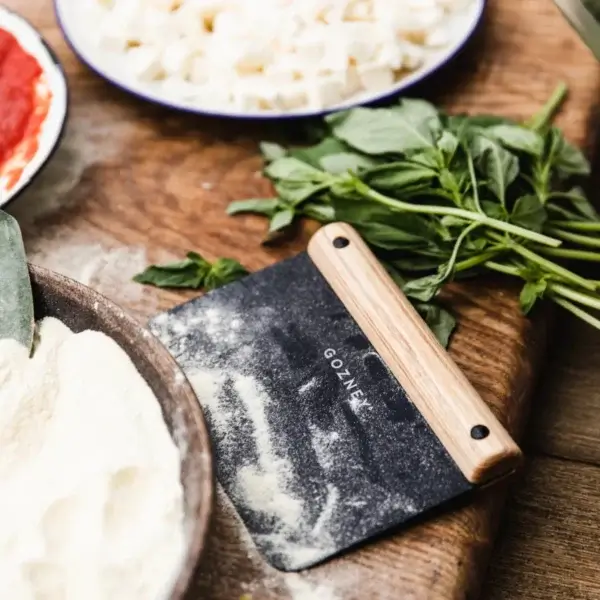 Gozney Dough Cutter con harina sobre la hoja, junto a albahaca, mozzarella y un bol de harina sobre mesa de madera.
