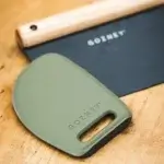 Primer plano de un raspador verde sobre mesa de madera con la Gozney Dough Cutter desenfocada al fondo.