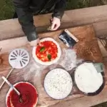 Preparación de pizza sobre mesa de madera con manos colocando albahaca, masa con tomate, mozzarella, harina y Gozney Dough Cutter.