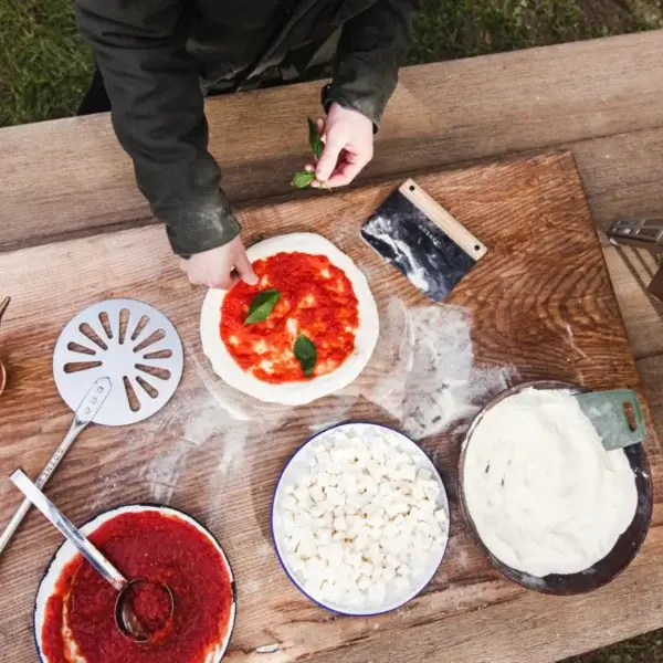 Preparación de pizza sobre mesa de madera con manos colocando albahaca, masa con tomate, mozzarella, harina y Gozney Dough Cutter.