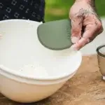 Mano con harina sujetando el Gozney Dough Scraper sobre un bol blanco con restos de masa.